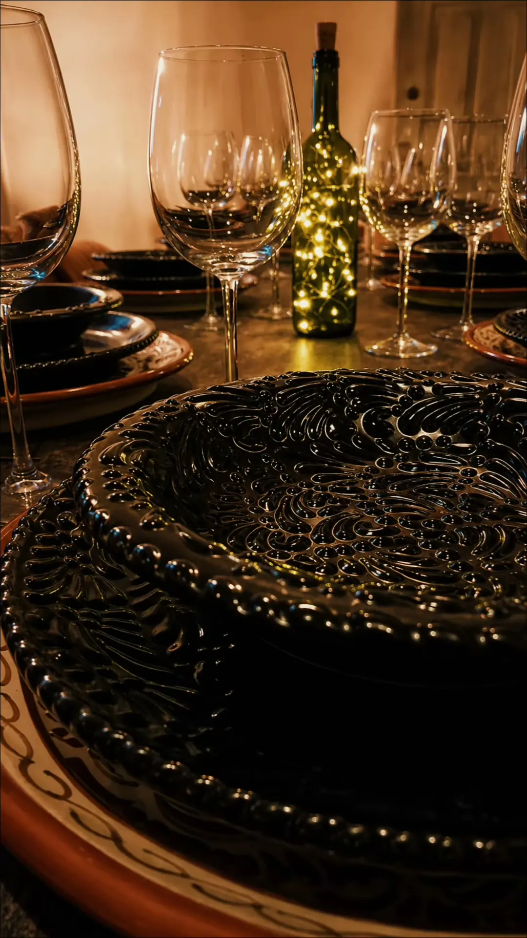 Mesa puesta en Piccola Regina: vajilla negra en relieve sobre platos de talavera, copas de vino y una botella con luces cálidas.