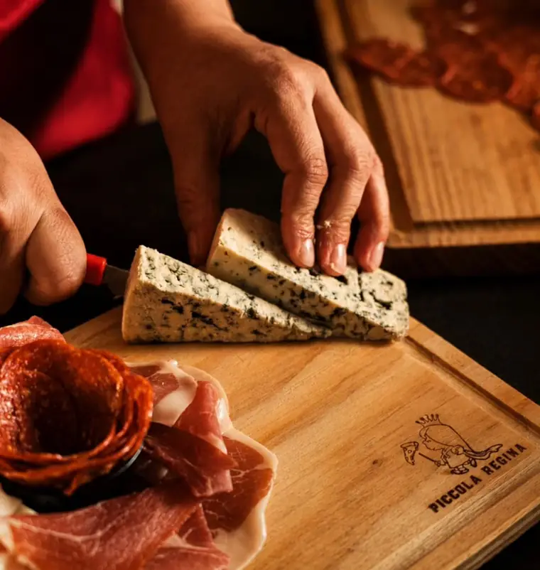Manos cortando queso azul sobre una tabla de madera grabada con el logotipo de Piccola Regina.